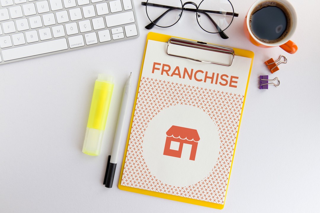 Clipboard on a desk labeled “FRANCHISE,” surrounded by office supplies including pens, binder clips, and a keyboard in a workspace setting.