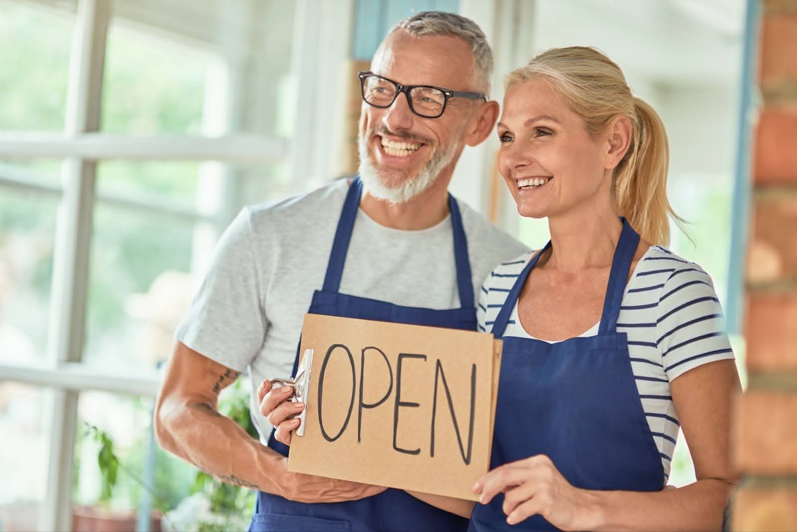 Older couple smiling holding up an "open" sign. 